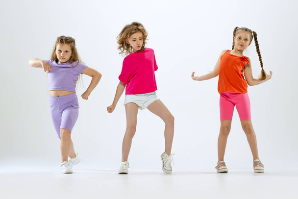 Dance group. Three little girls, kids in bright colorful clothes dancing, posing and jumping isolated on white studio background. Concept of music, fashion, art, childhood, hobby. Copy space for ad - Фото, изображение
