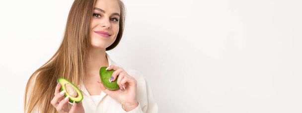 Portrait of a lovely smiling young brunette caucasian woman wearing the white shirt with long hair holding and showing avocado, standing isolated over white background - Zdjęcie, obraz