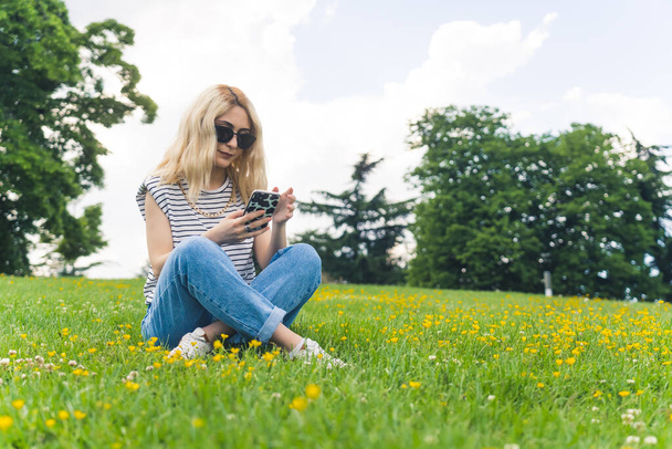 dość kaukaska dziewczyna w luźnych ubraniach i okularach przeciwsłonecznych siedzi na trawie i za pomocą telefonu, pełny strzał w parku. Wysokiej jakości zdjęcie - Zdjęcie, obraz