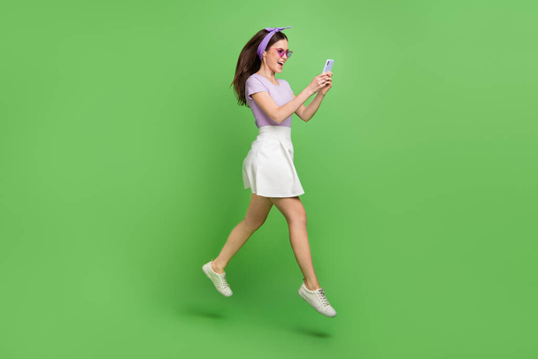 Full body photo of impressed brunette lady run look telephone wear head band glasses t-shirt skirt shoes isolated on green background. - Φωτογραφία, εικόνα
