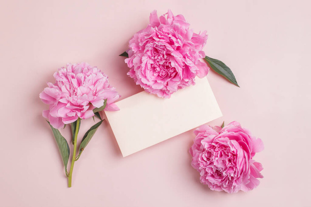 Tarjeta de invitación o felicitación maqueta con sobre y delicadas peonías rosadas sobre fondo rosa pastel. Vista superior - Foto, Imagen