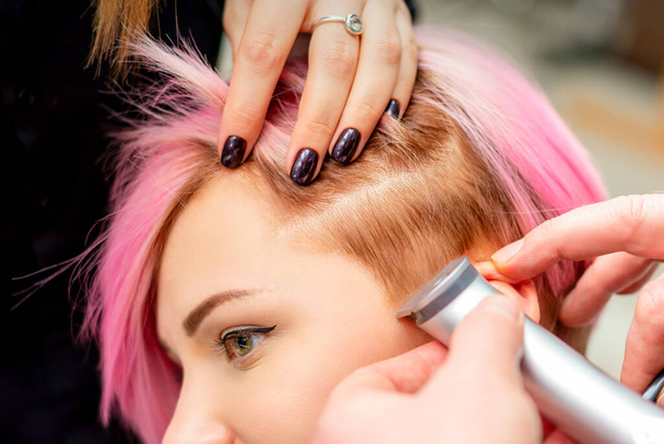 Парикмахер бреет женский храм с розовыми волосами электробритвой в парикмахерской, закрывается - Фото, изображение