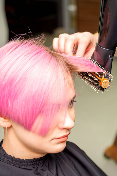 Séchage cheveux courts roses de jeune femme caucasienne avec un sèche-cheveux noir et brosse ronde noire par les mains d'un coiffeur masculin dans un salon de coiffure, gros plan - Photo, image