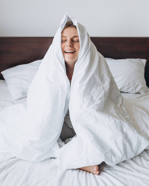 Beautiful young happy woman wrapped in soft white blanket sitting at home. Winter atmosphere. Millennial Caucasian girl wake up in cozy bed at home - Photo, Image