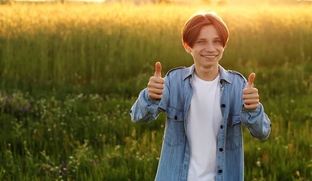Pozitivní teen mladý muž s úsměvem do kamery a ukazující palec nahoru ceduli venku při západu slunce. Koncept mládeže a pozitivních emocí - Fotografie, Obrázek