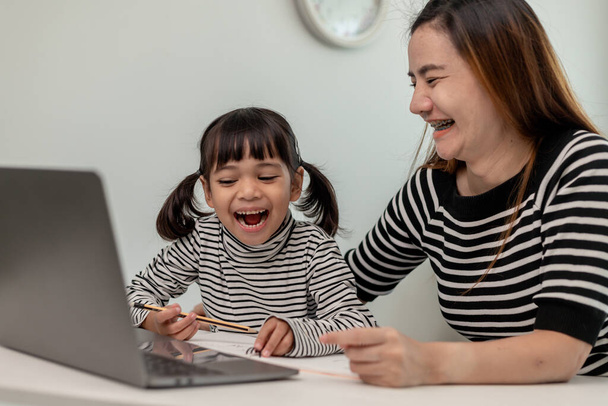 Asyalı küçük kız evde annesiyle online ders alıyor. Anaokulu çocuğu dizüstü bilgisayar kullanır ev ödevi yapar, okul öğretmeninden dijital uzaktan internete geçer ve annesinden destek alır.. - Fotoğraf, Görsel