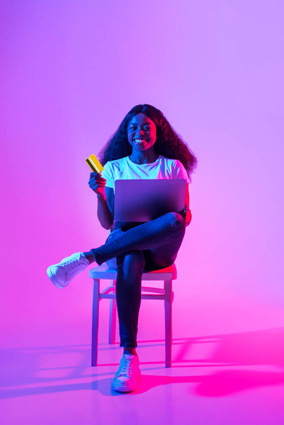 Online payments. Happy African American woman with credit card and laptop sitting on chair in neon light. Joyful young black female enjoying remote shopping or paying bills in internet - Photo, Image