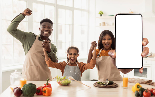 Happy Excited Black Family In Kitchen Pokazuje pusty smartfon i świętuje sukces, afroamerykańskich rodziców i córkę w fartuchach Pokazuje przestrzeń na aplikacje mobilne, kolaż, makietę - Zdjęcie, obraz