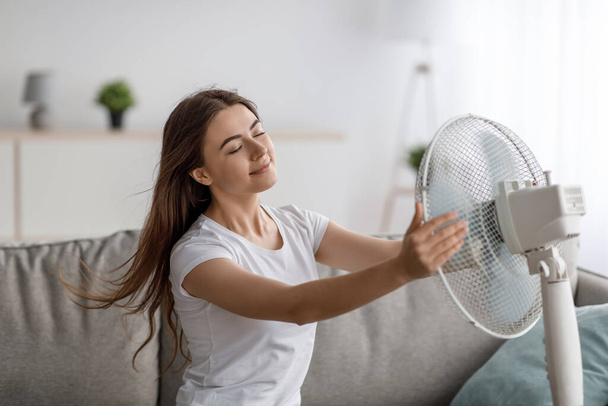 Satisfied pretty millennial caucasian female suffering from heat, sitting on sofa and enjoying cold air from fan in living room interior. Overheating without air conditioning, summer weather at home - Photo, image