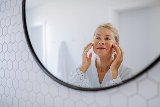 A beautiful senior woman in bathrobe, applying eye patches for puffiness while looking in the mirror - Photo, Image