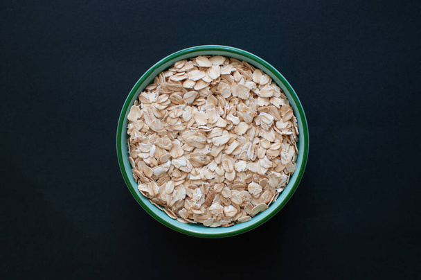 Top view of natural and nutritious oat flakes in green bowl. Popular cereal food isolated on dark background, selective focus. - Fotó, kép