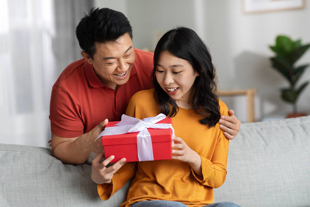 Loving asian man embracing from behind and giving gift box his beautiful young wife, making great surprise for St. Valentines Day or birthday, home interior, free space. Anniversary celebration - Photo, Image