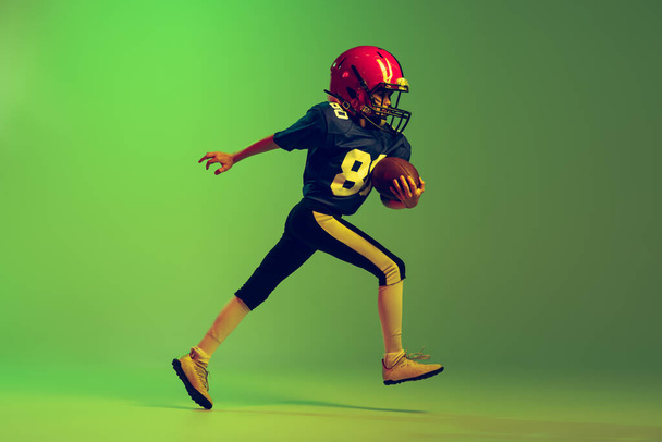 Running forward. Sportive little boy in sports uniform and equipment playing american football isolated on green background in neon light. Concept of sport, ad, movement, achievements. - Φωτογραφία, εικόνα