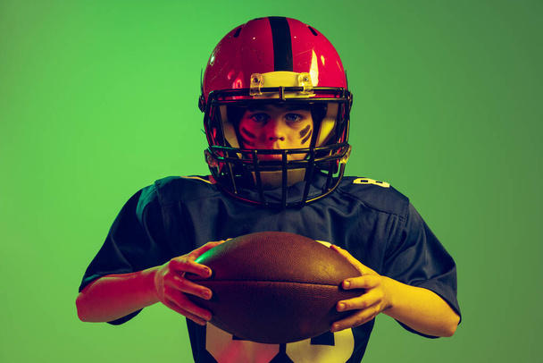 Setting to play and win. Closeup portrait of emotional boy, beginner american football player in sports uniform and helmet isolated on green background in neon light. Concept of sport, challenges - Фото, изображение