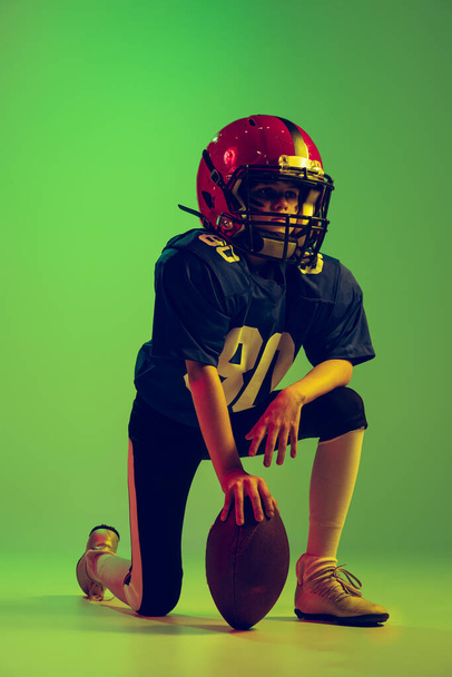 Starting position. Athletic kid, beginner american football player in sports uniform and helmet training isolated on green background in neon. Concept of sport, challenges, action, achievements. - 写真・画像