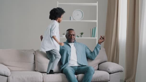 Mature tired man sitting on sofa in room meditating funny active little daughter jumping on couch near dad noisy kid girl prevents father from relaxing bad behavior disobedience parenthood concept - Footage, Video