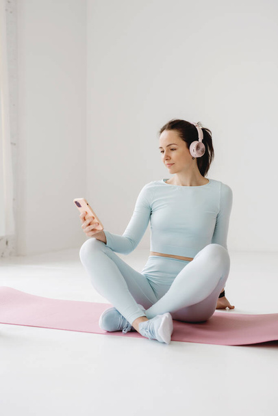 Young woman in blue sportswear and pink headphones holding and using smartphone sitting on yoga mat. - Fotoğraf, Görsel