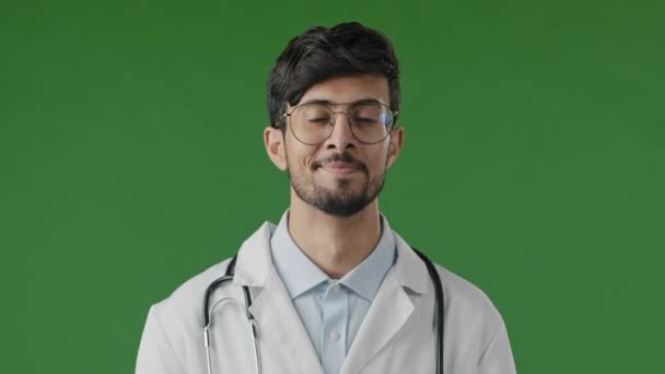 Portrait of smiling arab guy pharmacist therapist professional physician doctor wears white coat with stethoscope look at camera male medical adviser stand posing isolated in green studio background - Footage, Video