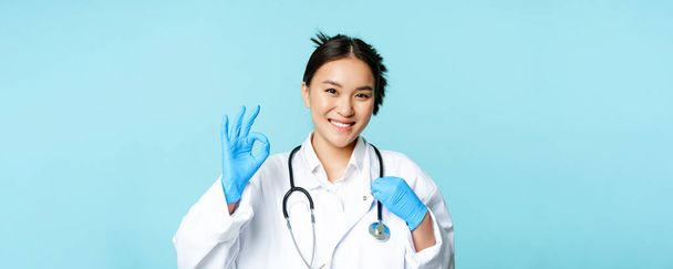Smiling asian doctor, female physician touching stethoscope, showing okay, ok sign in approval, blue background. - Фото, зображення
