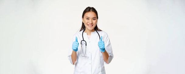 Sorridente medico asiatico, donna in uniforme medica e guanti di gomma mostra pollici in su, raccomandando smth al paziente, in piedi su sfondo bianco. - Foto, immagini