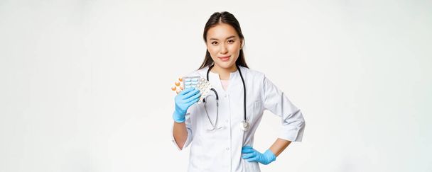 Asian female physician showing pills, medication, holding vitamins and wearing rubber gloves, standing over white background. - Photo, image