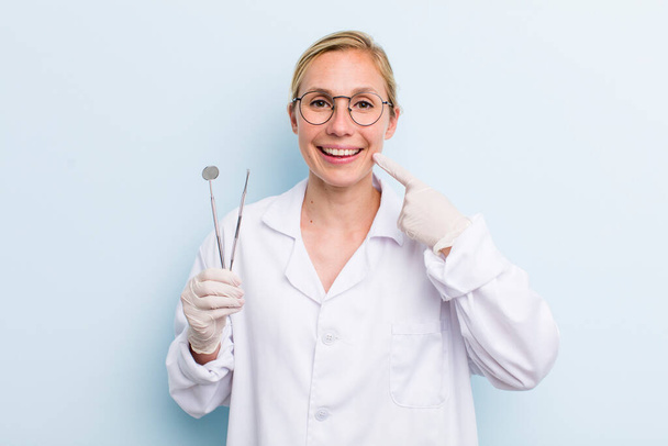young adult blonde woman smiling confidently pointing to own broad smile. dentist concept - Foto, Bild