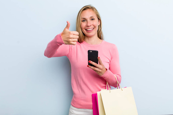 young adult blonde woman feeling proud,smiling positively with thumbs up. shopping bags concept - Photo, Image