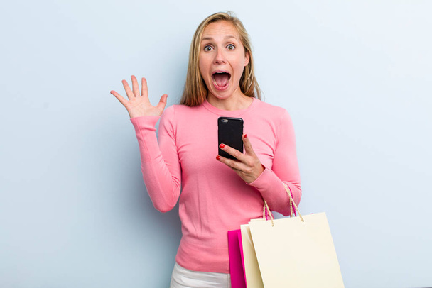 young adult blonde woman feeling happy and astonished at something unbelievable. shopping bags concept - Photo, Image