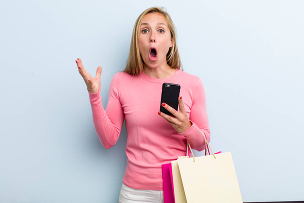 young adult blonde woman feeling extremely shocked and surprised. shopping bags concept - Foto, Imagem