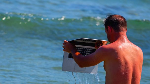 あなたのラップトップコンピュータの世話をする時間。海によって夏休みに男の面白いユーモラスなイメージは、水の中で彼のラップトップを洗う.  - 写真・画像