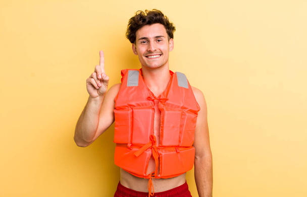 young handsome guy smiling and looking friendly, showing number one. life jacket and boat concept - Zdjęcie, obraz