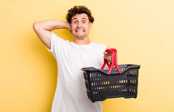 young handsome guy feeling stressed, anxious or scared, with hands on head. empty shopping basket concept - Фото, изображение