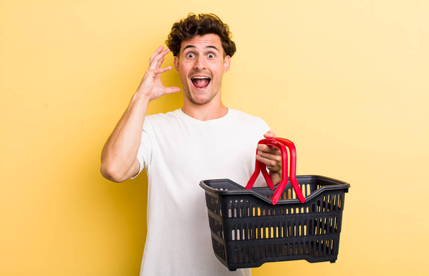 young handsome guy screaming with hands up in the air. empty shopping basket concept - Фото, изображение