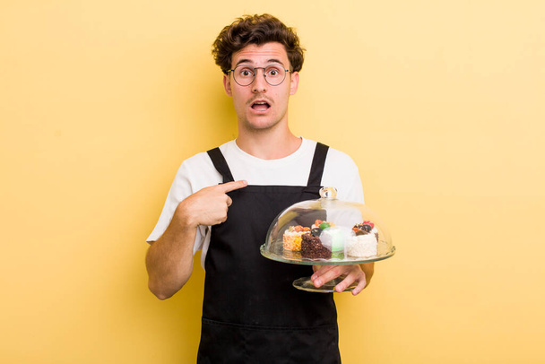 young handsome guy looking shocked and surprised with mouth wide open, pointing to self. home made cakes concept - Fotoğraf, Görsel