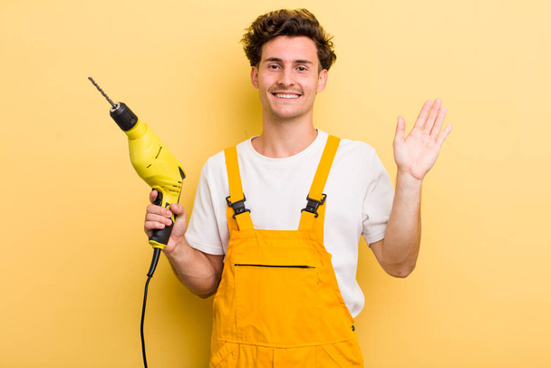 young handsome guy smiling happily, waving hand, welcoming and greeting you. handyman with a drill concept - Фото, изображение