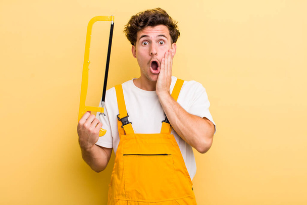 young handsome guy feeling shocked and scared. handyman and a saw concept - Fotó, kép