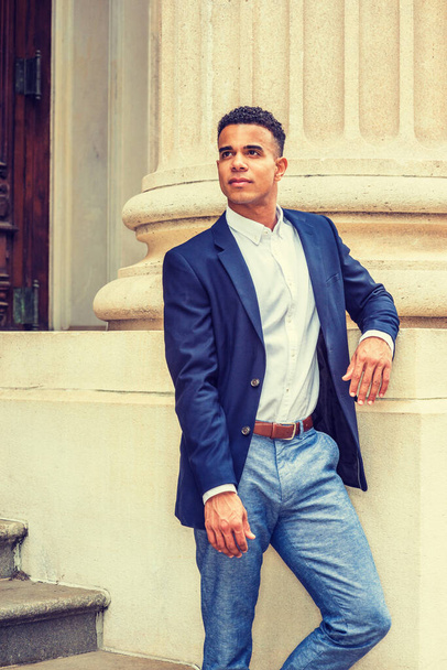Empresario afroamericano trabajando en Nueva York. Usando chaqueta azul, camisa blanca, pantalones grises, un estudiante universitario negro parado por la columna afuera en el campus, mirando hacia arriba. - Foto, Imagen
