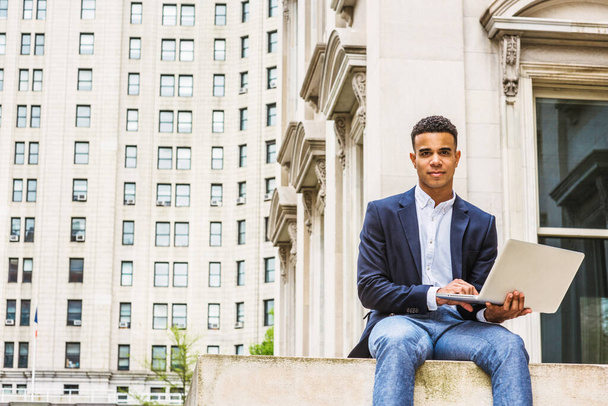 Afro-Amerikaanse student studeert in New York, draagt blauwe blazer, wit shirt, grijze broek, zit op straat buiten kantoorgebouw op de campus, werkt aan laptop computer, denkt na - Foto, afbeelding