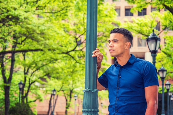 Afroamerikanische College-Studentin, trägt blaues Kurzarmshirt, steht im Sommer an einem Lichtmast auf der Straße auf dem Campus in New York, schaut weg, denkt nach, verliert sich in Gedanken.  - Foto, Bild