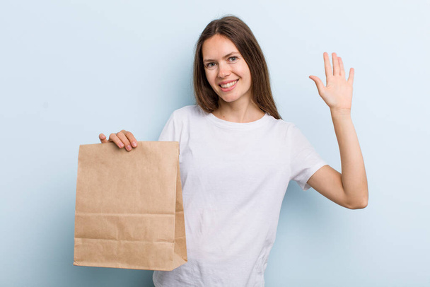 young adult woman smiling happily, waving hand, welcoming and greeting you. delivery and take away concept - Foto, Imagem