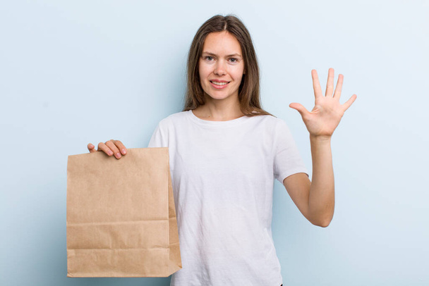 young adult woman smiling and looking friendly, showing number five. delivery and take away concept - Foto, afbeelding