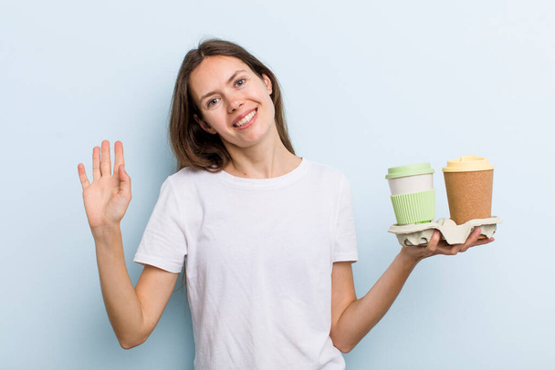 young adult woman smiling happily, waving hand, welcoming and greeting you. take away coffee concept - Фото, изображение