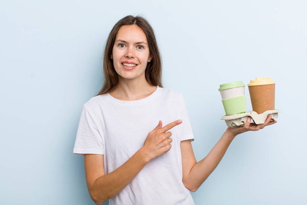 young adult woman smiling cheerfully, feeling happy and pointing to the side. take away coffee concept - Фото, изображение