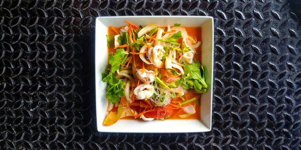 Top view of Thai spicy seafood salad with glass noodle, squid, shrimp, sliced tomato, onion, carrot and celery on white dish or plate. Flat lay of Asian food on black stainless steel background. - Photo, Image