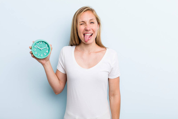 blonde young adult woman with cheerful and rebellious attitude, joking and sticking tongue out. alarm clock concept - Fotoğraf, Görsel