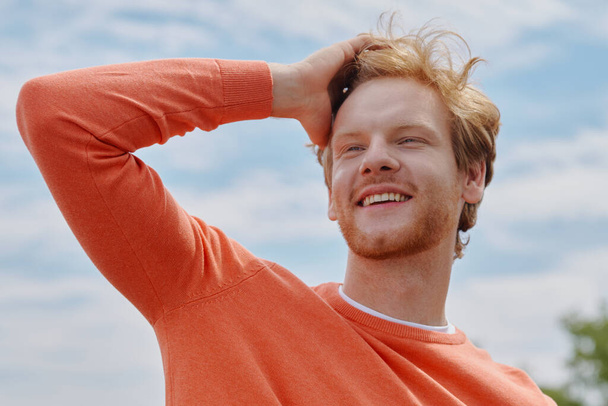 Happy young redhead man holding hand in hair and smiling while standing outdoors - Фото, изображение
