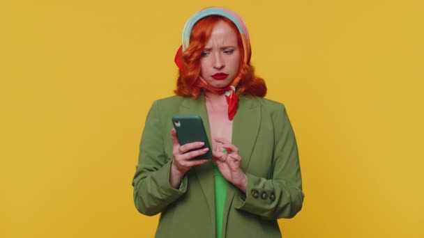 Разочарованная рыжая молодая женщина использует смартфон, печатающий в браузере, теряет удивление внезапными результатами лотереи, плохими новостями удачи, потерей. Рыжая девушка в помещении студии изолированы на желтом фоне - Кадры, видео