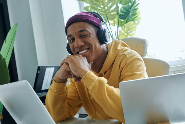 Alegre hombre de raza mixta en auriculares sentados en su lugar de trabajo y guiñando un ojo - Foto, Imagen