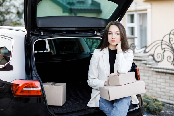 Menina sentada na bagageira do carro, segurando caixas em suas mãos e sorrindo, olhando para a câmera - Foto, Imagem