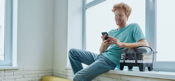Cheerful redhead man using smart phone while sitting on the window sill at home - Fotoğraf, Görsel